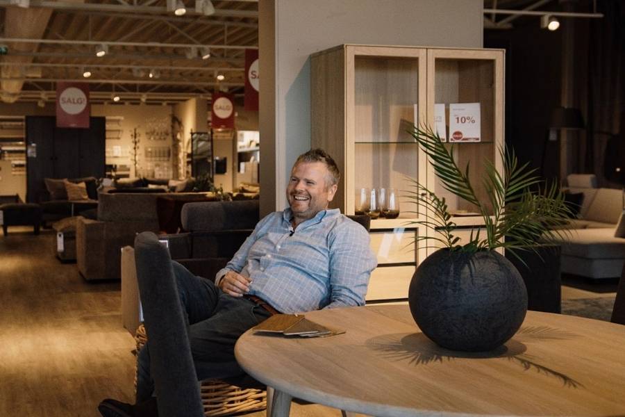 Person sitting in a furniture store on a chair with legs crossed, holding a drink. The face of the person is blurred out. In the foreground, there is a round wooden table with an open book and a large black vase containing green plants. Behind the person, various pieces of furniture including sofas, chairs, and shelves are visible. Several sale signs can be seen in the background indicating items are on sale.