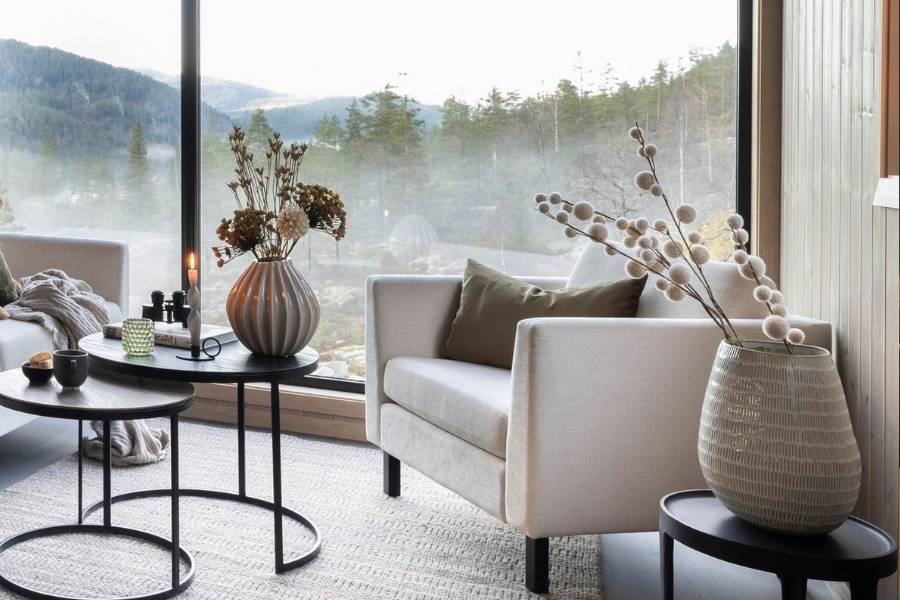 Modern living room with a large window offering a scenic view of mountains and trees. The room features a light-colored sofa with green and beige cushions, an armchair on the right, and two round, black metal coffee tables with decorative items like a vase with dried flowers, candles, and small bowls. The floor has a textured rug in neutral tones, and the walls are made of light wood paneling.